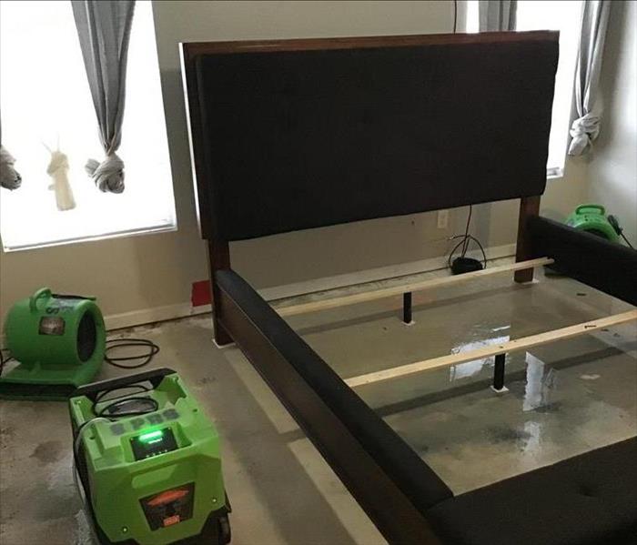 Professional SERVPRO green air movers and dehumidifiers are placed in a bedroom around a bed frame with water on the ground